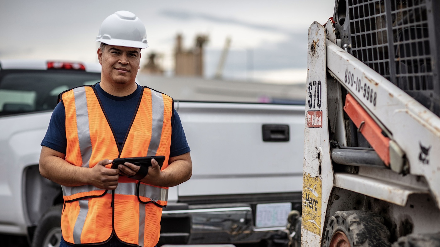 man in construction hard hat standing next to a bobcat on a truck trailer